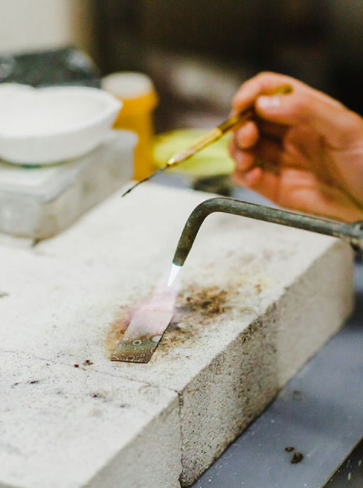 A strip of metal resting on a block of concrete under a torch which is heating the material. Also there is a hand holding an thin long tool relating to the work.