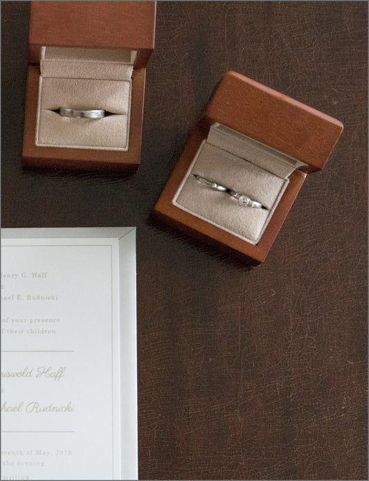 On a dark wooden table there is a wedding invitation and 2 wooden boxes. One wooden box contains a silver band and the other has a silver band and a silver diamond ring.