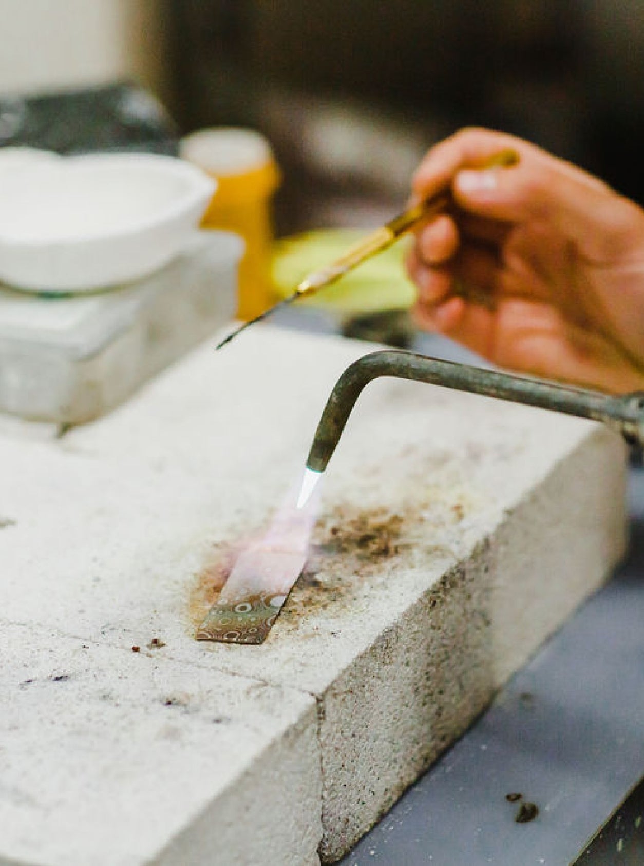 A strip of metal resting on a block of concrete under a torch which is heating the material. Also there is a hand holding an thin long tool relating to the work. 