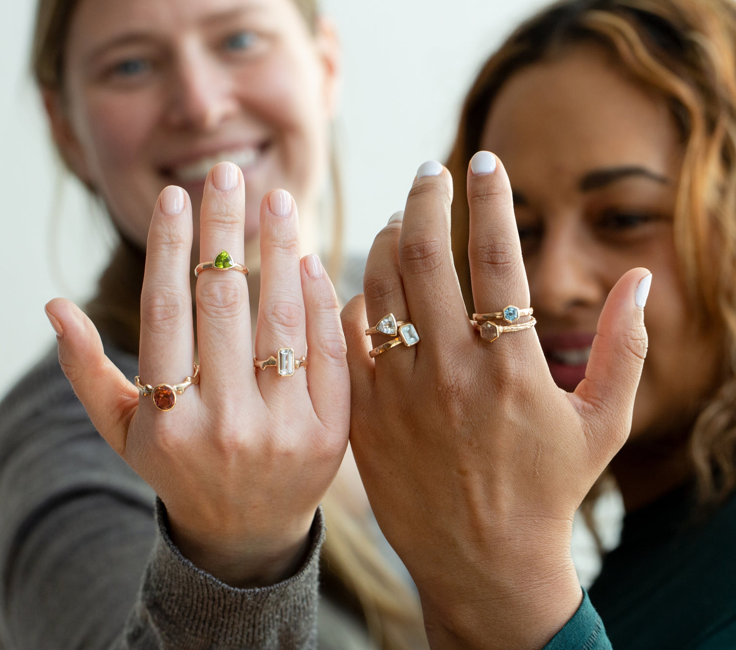 A blonde haired and brown haired woman each holding up their left hands. On their hands are various rings handcrafted by W.R. Metalarts from the Gold for Good collection which are made with 100% Fairminded gold. This gold is certifiably sourced.