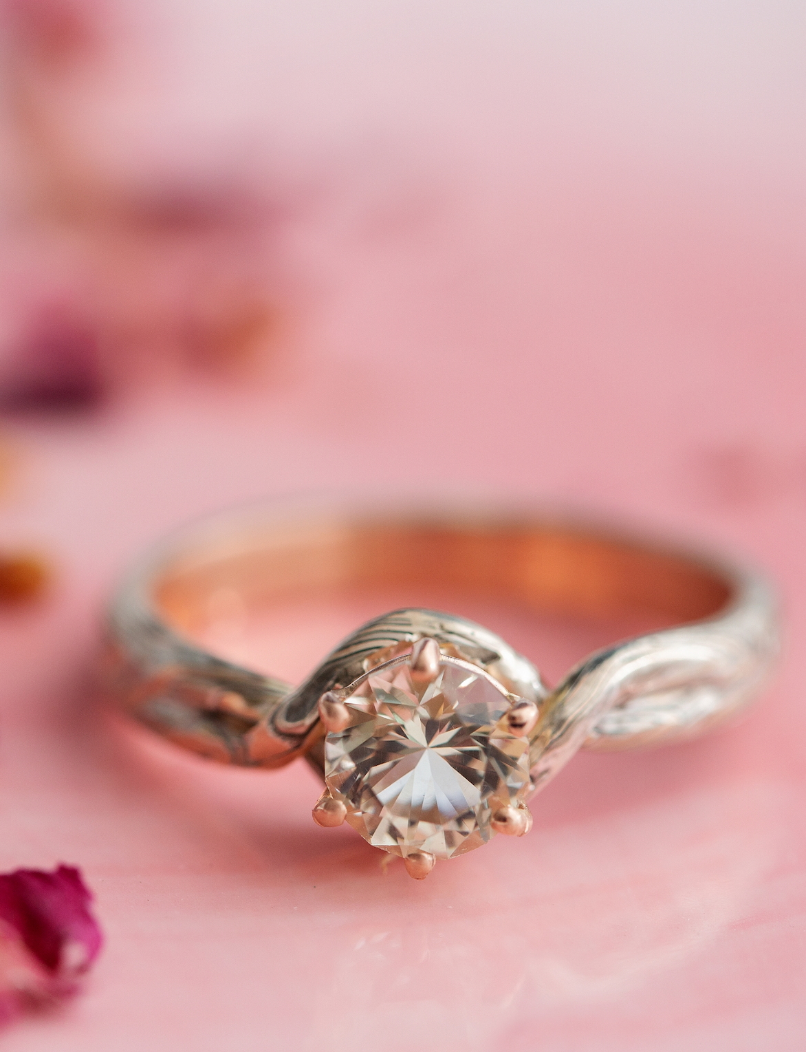 A ring made of rose gold mokume gane made of layers of rose gold, white gold, and silver placed on a pink background surrounded with rose petals